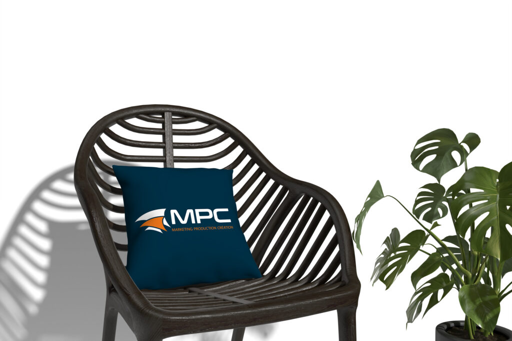 Coussin carré bleu marine personnalisé avec le logo MPC, disposé sur un fauteuil en rotin foncé à côté d'une plante Monstera sur fond blanc.