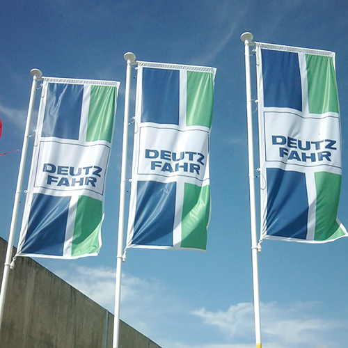 Trois drapeaux publicitaires Deutz-Fahr flottant sur des mâts sous un ciel bleu.