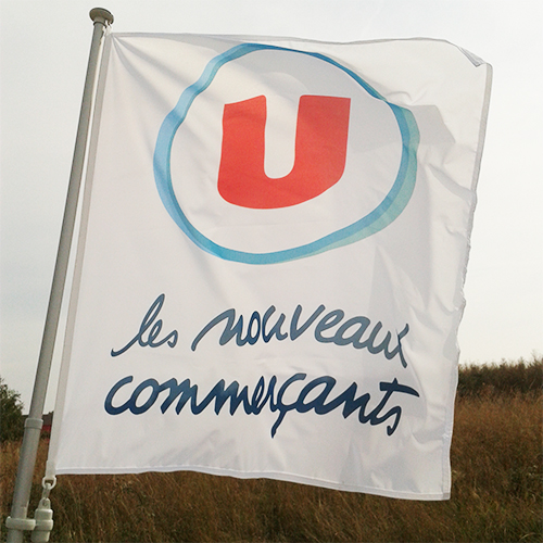 Drapeau publicitaire blanc de l'enseigne Système U flottant au vent sur un mât en aluminium, affichant le logo "U" rouge et le slogan "les nouveaux commerçants".