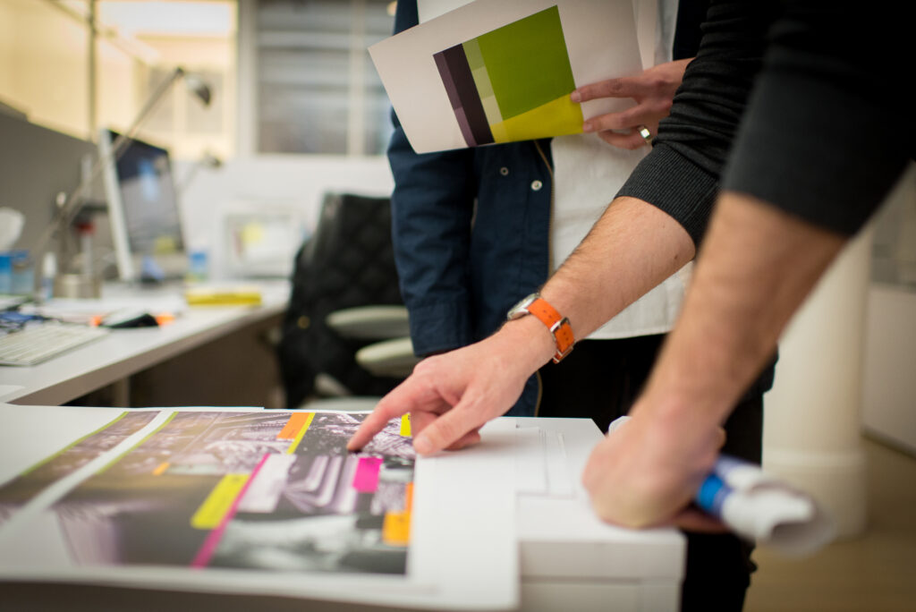 Deux professionnels travaillent sur des supports de communication imprimés dans un bureau, en examinant des maquettes graphiques et des échantillons de couleurs.